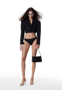 Czarny przycięty żakiet na czarnym bikini, w komplecie z małą czarną torebką i białymi sandałkami na obcasie z paskami. Długie, falowane włosy i niebieskie okulary przeciwsłoneczne.