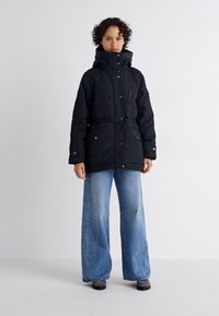 Svart vadderad parka-jacka med hög krage, dragsko i midjan och framfickor. Kombinerad med vida ljusblå jeans och svarta skor.
