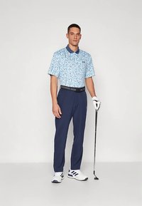 Mies, joka on pukeutunut siniseen kuvioituun poolopaitaan, tummansinisiin housuihin, valkoiseen golfkäsineeseen ja lenkkareihin, pitelee golfmailaa ja seisoo yksiväristä taustaa vasten.