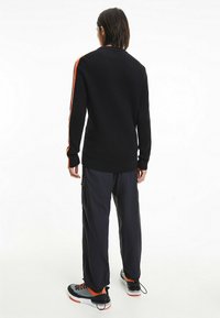 Schwarzer gerippter Pullover mit orangen Seitenstreifen, kombiniert mit locker sitzenden marineblauen Cargo-Hosen und schwarzen Sneakers mit roten Akzenten.