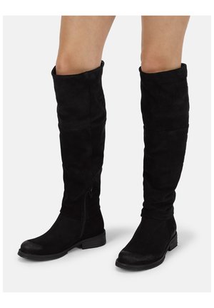 Schwarze kniehohe Wildlederstiefel mit niedrigen Absätzen und seitlichen Reißverschlüssen, getragen auf nackten Beinen vor weißem Hintergrund.