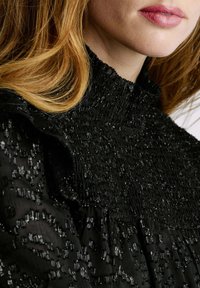 Blusa nera con texture, dotata di un alto collo arricciato, caratterizzata da motivi in filo lucido e maniche con rouches, che mostra un design a strati.