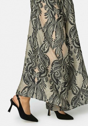 Persoană purtând pantofi negri cu toc înalt, model slingback, și o fustă lungă, transparentă, cu un model negru floral și de frunze peste un material nud.