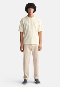 Cremfarbendes Baumwoll-T-Shirt, lockere Passform, kurze Ärmel. Beigefarbene Hose, gerader Schnitt. Blaue Sneakers mit weißen Akzenten und Gummisohle, lässig getragen.