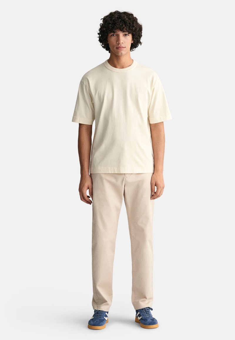 Cremfarbendes Baumwoll-T-Shirt, lockere Passform, kurze Ärmel. Beigefarbene Hose, gerader Schnitt. Blaue Sneakers mit weißen Akzenten und Gummisohle, lässig getragen.