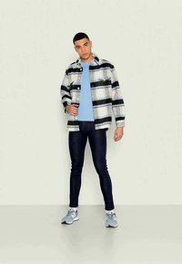 Casaco xadrez em preto, branco e cinza, usado sobre uma t-shirt azul clara. Jeans escuros e ténis cinzentos completam o visual. Postura em pé.