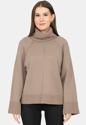 Mujer que lleva un suéter de cuello alto color topo con puños anchos y pantalones negros, de pie frente a un fondo liso.