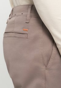 Lichtbruine broek van een gladde stof met een achterzak met een klein oranje "BOSS" logo. De riemlussen en zijzakken zijn zichtbaar.