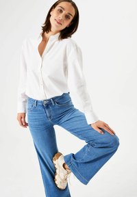 Frau in weißem Hemd und blauen Jeans, die ein Bein hebt und beigen Sneakern trägt, vor einem schlichten weißen Hintergrund.