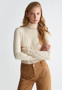 Maglione a collo alto di colore crema con texture a coste, caratterizzato da un scollo ondulato e dettagli con bottoni sulle maniche. Abbinato a pantaloni marroni con tasche chiodate.