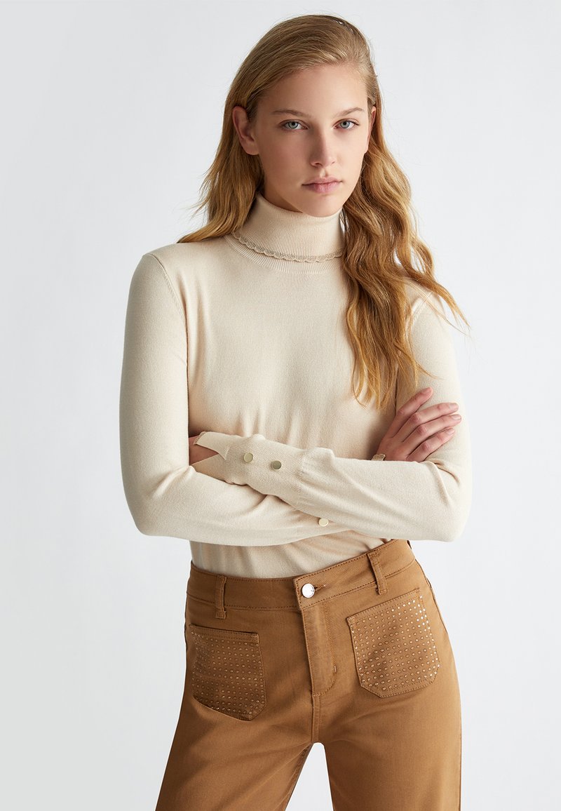 Maglione a collo alto di colore crema con texture a coste, caratterizzato da un scollo ondulato e dettagli con bottoni sulle maniche. Abbinato a pantaloni marroni con tasche chiodate.