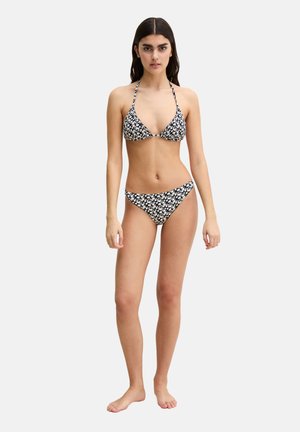 Kobieta stojąca boso w czarno-białym bikini w kwiatowy wzór na wiązanym dekolcie na jednolitym białym tle.
