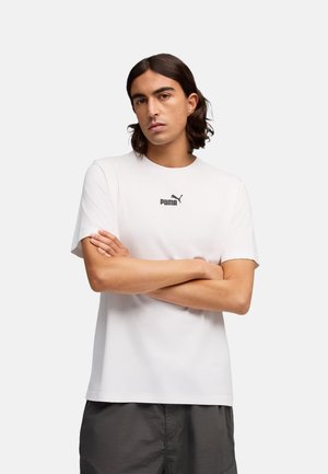 Młody mężczyzna o włosach do ramion, ubrany w białą koszulkę Puma, z założonymi skrzyżowanymi rękoma na jasnym, jednolitym tle.