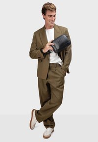 Olivenfarbener Anzug mit einem weißen T-Shirt, hält eine schwarze Clutch-Tasche. Sneakers mit weißem Mesh und braunen Akzenten. Modern, taillierte Passform.