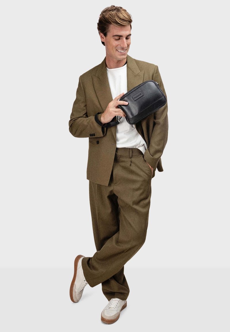 Olivenfarbener Anzug mit einem weißen T-Shirt, hält eine schwarze Clutch-Tasche. Sneakers mit weißem Mesh und braunen Akzenten. Modern, taillierte Passform.