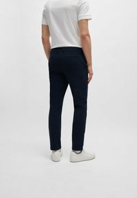 Persoon in een marineblauwe slim-fit broek, witte sneakers, witte enkelsokken en een witte T-shirt met korte mouwen, staand tegen een witte achtergrond.
