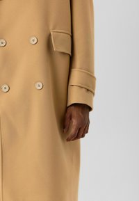 Beige double-breasted jas met een zachte textuur, grote knopen en een zijzak, met een detail van omgeslagen mouwen.