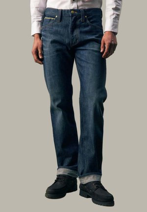 Mann trägt dunkelblaue Jeans mit hochgekrempelten Säumen, schwarze Schnürschuhe und ein weißes Langarmhemd, steht vor einem schlichten Hintergrund.