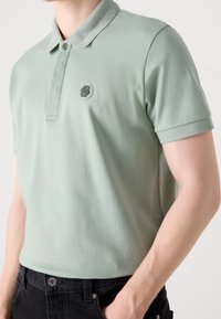 Camisa polo verde claro con cuello acanalado, mangas cortas y un logotipo bordado circular en el pecho. Tejido de algodón suave.