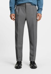 Grijze, op maat gemaakte broek met een gestructureerde afwerking, voorzien van plooien, zijzakken en een glad front, gecombineerd met zwarte instapschoenen.