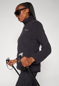 Black fleece jacket with high collar, zip closure, and two side pockets. Model holds poles with gray grips; smooth fabric texture.