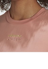 Světle růžové tričko s krátkým rukávem vyrobené z jemné tkaniny. Má vyšívané zlaté logo "adidas" a akcent listu na hrudi. Hladká textura.
