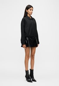 Μαύρο μπουφάν bomber με γιακά και κλείσιμο με φερμουάρ, συνδυασμένο με μαύρη γυαλιστερή πλισέ φούστα και μαύρα, ψηλοτάκουνα μποτάκια αστραγάλου.