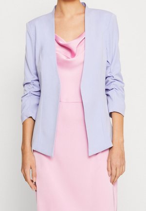 Femme portant un blazer lavande avec des manches froncées sur une robe rose en satin à col bénitier, debout devant un fond blanc uni.
