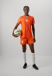 Oranžový futbalový dres Nike s textúrovaným tričkom a šortkami, ktorý má emblém leva. Hráč drží mnohobarebnú futbalovú loptu a má na sebe biele ponožky a kopačky.