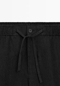 Bukser med sort elastik i taljen, bindebånd og enkelt sort knaplukning foran i midten.