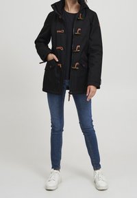 Schwarze wasserdichte Jacke mit Kapuze, ausgestattet mit braunen Kordelverschlüssen, zwei Fronttaschen und einem Reißverschluss vorne. Kombiniert mit blauen Jeans und weißen Sneakern.