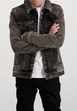 Persona con chaqueta de mezclilla negra descolorida sobre camiseta blanca con los brazos cruzados, combinada con pantalones negros, de pie frente a un fondo liso.