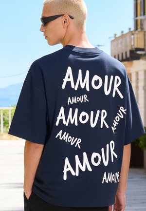 Jong persoon met kort blond haar en een zonnebril, gekleed in een oversized donkerblauw t-shirt met het woord "AMOUR" meerdere keren in het wit op de achterkant.