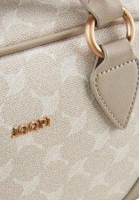 Beige stoffen handtas met een subtiele reliëfpatroon, goudkleurige hardware en een decoratieve leren accent met stikdetails.