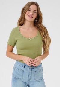Top verde de manga corta con detalle de encaje en el escote y lazo en el centro, combinado con jeans de tiro alto en azul claro.
