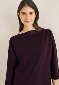 Ciemnofioletowa bluzka wykonana z miękkiej tkaniny, z szerokim dekoltem typu łódka i rękawami długości do połowy przedramienia, z delikatnym połyskiem na krawędziach.