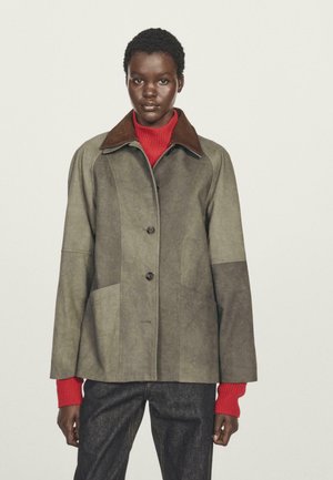 Femme noire avec les cheveux courts portant une veste en daim grise bicolore par-dessus un pull rouge vif et un pantalon foncé, se tenant devant un fond uni.