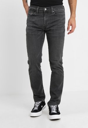 Mand iført slim-fit falmede sorte jeans og sorte lave sneakers med hvide snørebånd og sål, stående på en hvid gulv.