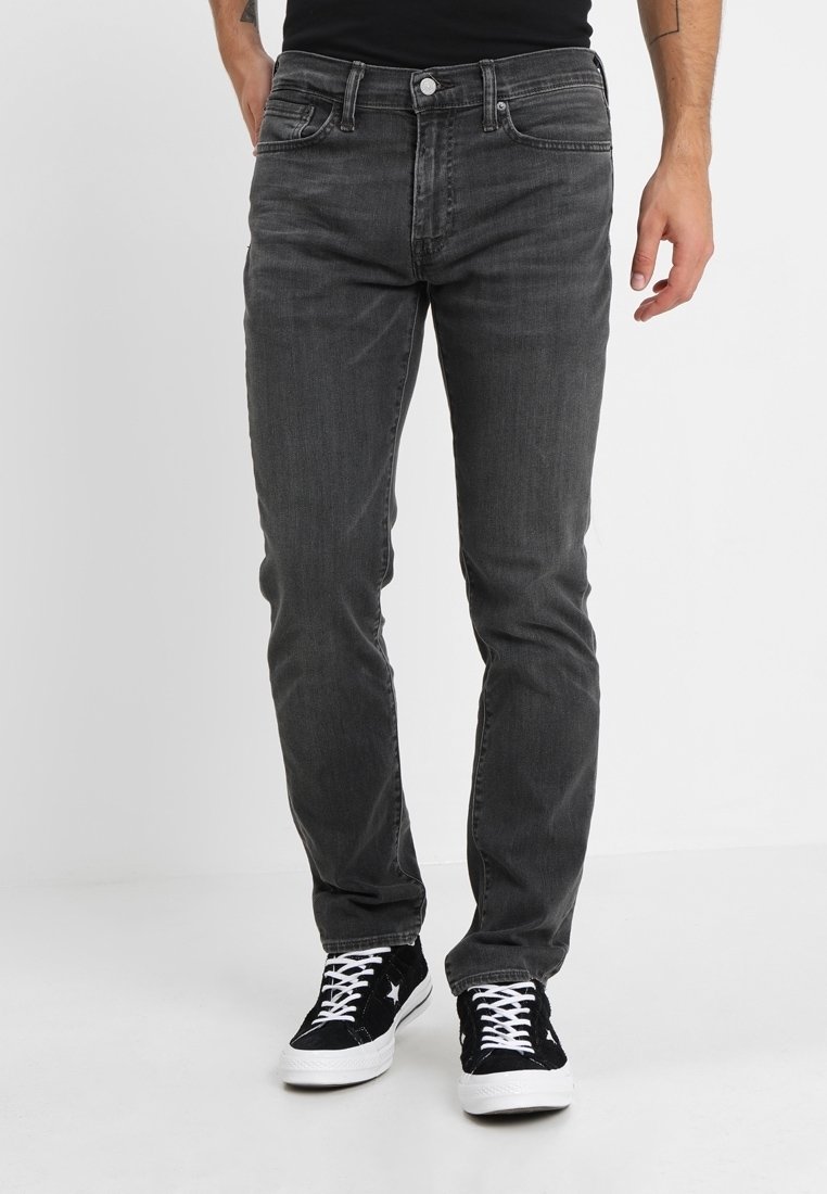 Homme portant un jean noir délavé slim et des baskets basses noires avec des lacets et une semelle blanches, debout sur un sol blanc.