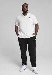 Polo bianco a maniche corte, logo nero sul petto, abbinato a pantaloni della tuta neri e sneakers bianche. Design sportivo semplice.