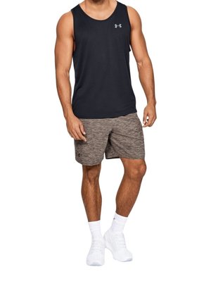 Hombre con camiseta deportiva sin mangas negra, pantalones cortos deportivos grises, calcetas blancas y zapatillas blancas, de pie contra un fondo blanco.