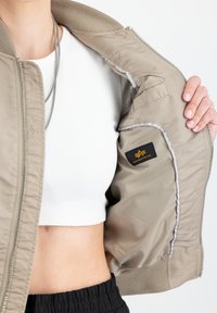 Béžová bomber bunda s ribovaným golierom a manžetami, so zipsom vpredu, prešívanou podšívkou a čiernou nášivkou s zlatým logom vo vnútri.