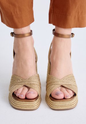 Pies con sandalias de plataforma tejidas en dorado con correas en el tobillo y pantalones cortos de color óxido sobre un fondo blanco.