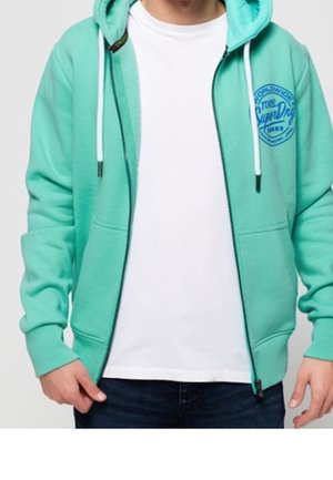 Man draagt een turquoise hoodie met rits en witte koordjes over een eenvoudige witte T-shirt en donkere jeans, handen ontspannen langs het lichaam.