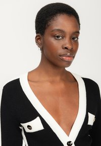 Schwarz-weißer Cardigan mit tiefem V-Ausschnitt, kontrastierenden Taschenakzenten und goldenen Knopfverzierungen, ausgestattet mit einem strukturierten Strickdesign.