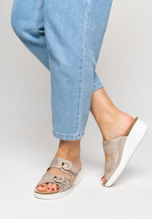 Feet wearing light gray slip-on wedge sandals with round buckles, paired with light blue rolled-up jeans, against a plain light background.