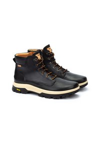 Botas de cuero negro hasta el tobillo con suela beige, que presentan cordones marrones, ojales de metal y una cremallera lateral. Suela de goma texturada para mayor agarre.