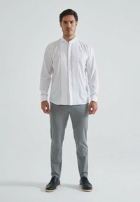 Homme portant une chemise plissée blanche à manches longues, un pantalon gris et des chaussures de ville noires, se tenant devant un fond lumineux et uni.