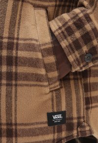Brązowa koszula flanelowa w kratę z miękką teksturą, z guzikami przy mankietach oraz metką Vans, z naprzemiennymi ciemnymi i jasnymi wzorami w kratkę.