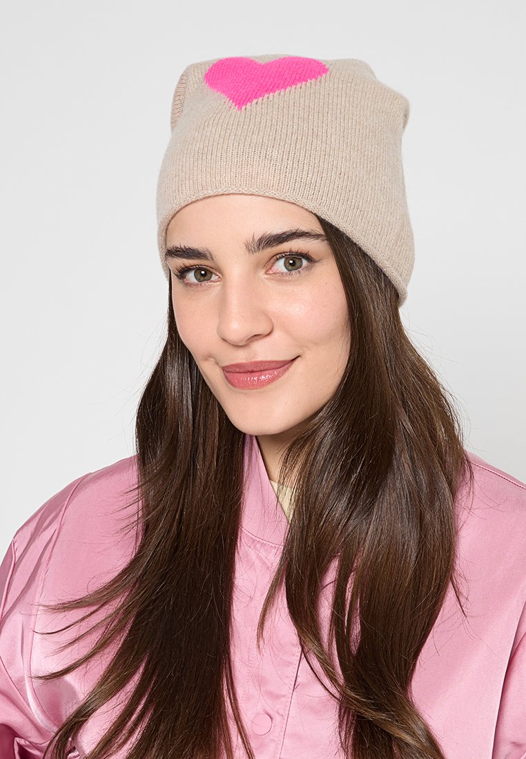 Beige gestrickte Mütze mit einem großen pinken Herzdesign auf der Vorderseite. Weicher Stoff und lässige Passform. Getragen mit einer pinken Satinjacke.